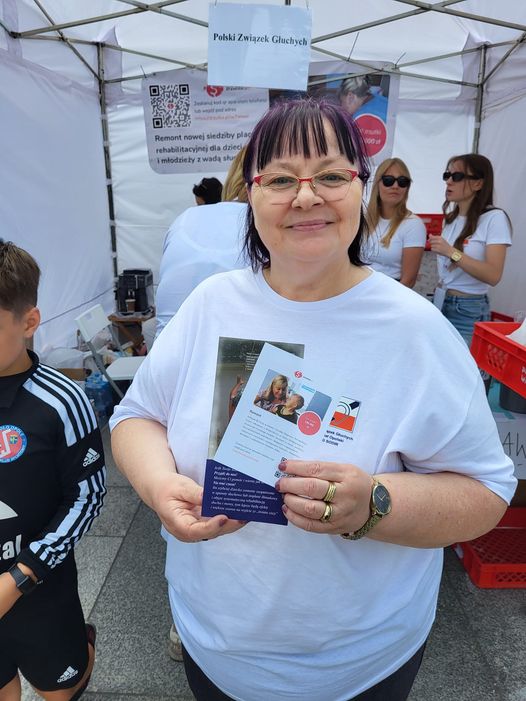 Na zdjęciu uśmiechnięta kobieta w ciemnych włosach i okularach. W rękach trzyma ulotki.